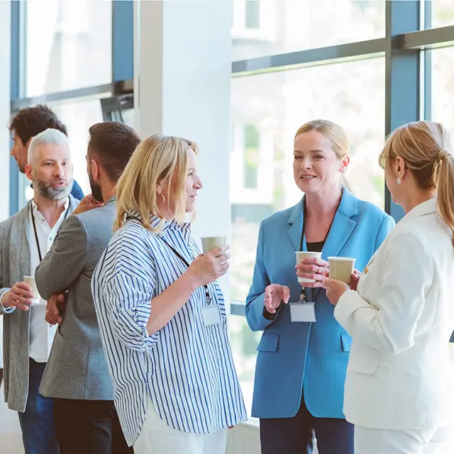 Kaffepause på konferanse, tre fornøyde kvinner i front av bildet, flere mennesker bak