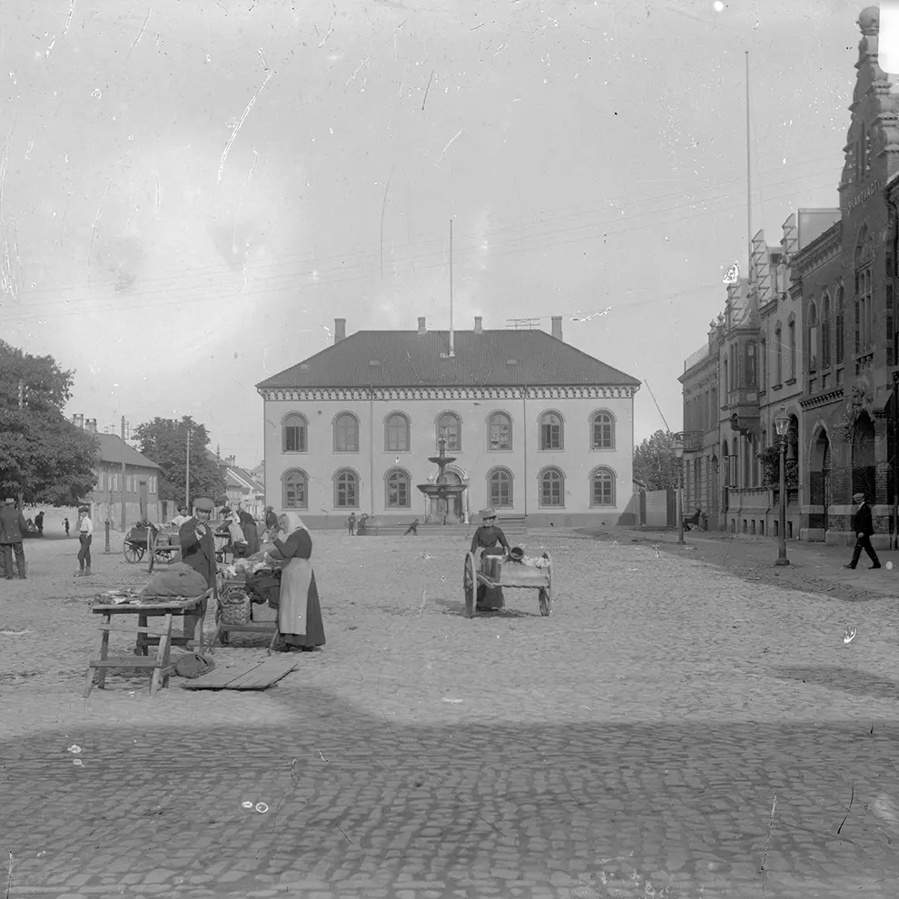 Historisk gatebilde fra Kristiansand med brostein, torghandel og eldre bygninger.