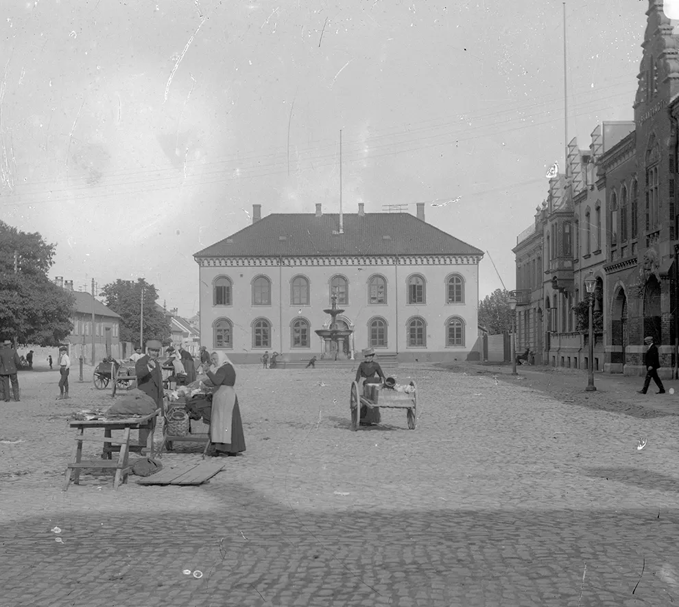 Historisk gatebilde fra Kristiansand med brostein, torghandel og eldre bygninger.