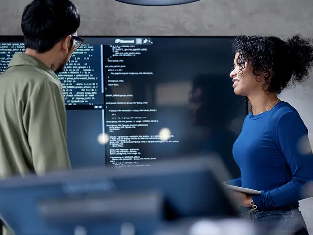 To utviklerkolleger diskuterer et prosjekt med skjermer rundt seg.