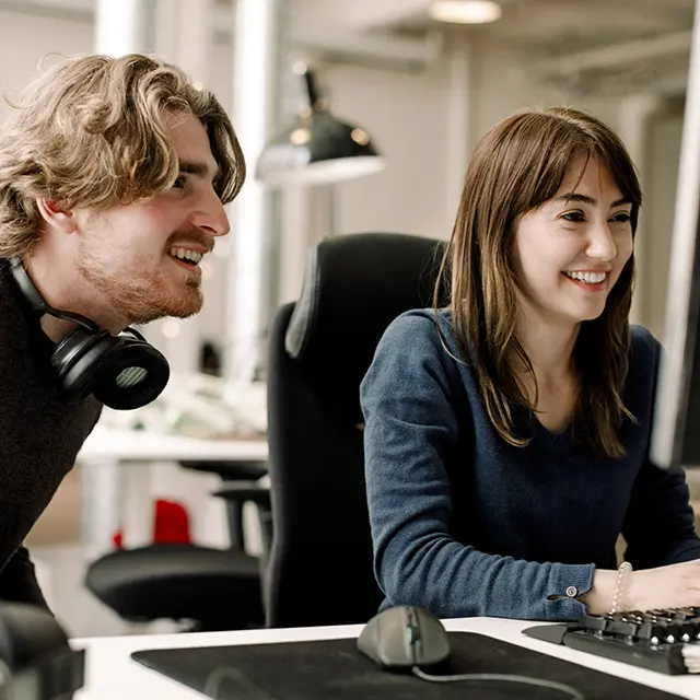 Mann og jvinne sitter ved kontorpult ser på skjerm smiler
