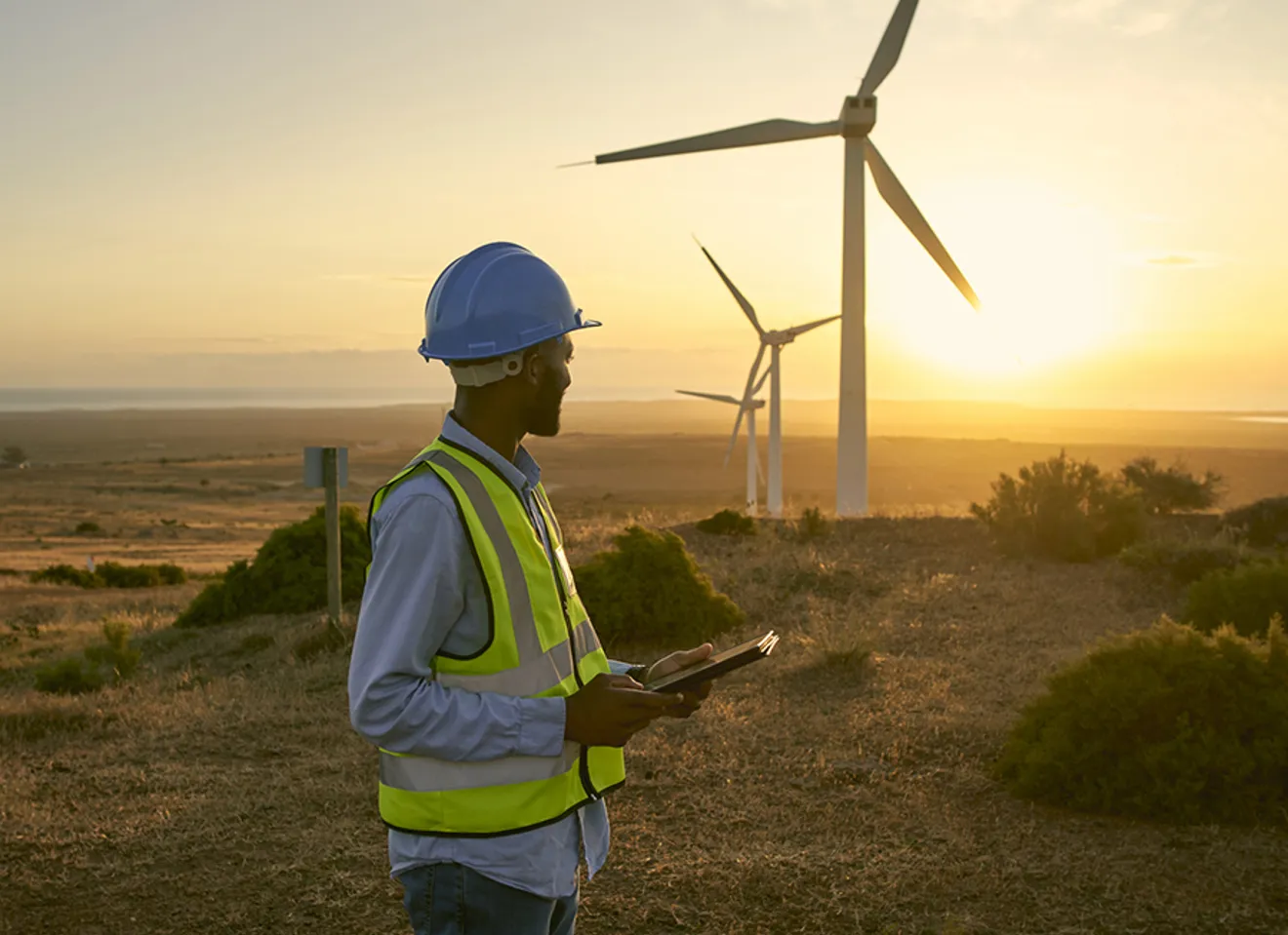 Ingeniør med nettbrett ved vindmøller i solnedgang på fornybar energiprosjekt.
