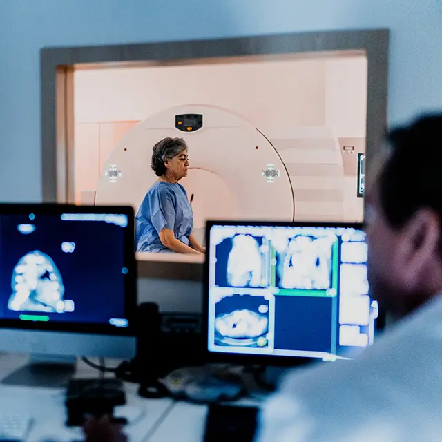 En eldre kvinne sitter i et rom for prøvetaking, lege og skjermer med prøveresultater i forgrunnen.