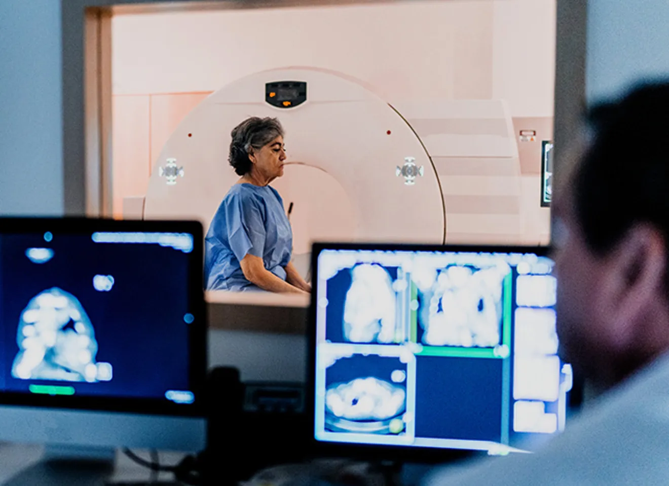 En eldre kvinne sitter i et rom for prøvetaking, lege og skjermer med prøveresultater i forgrunnen.