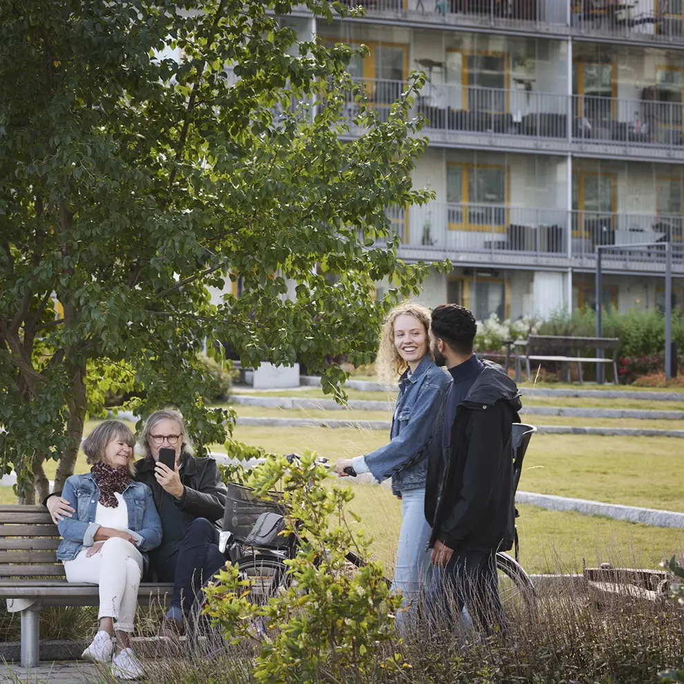 Et eldre par på benk og et ungt par med sykkel i uteområdet til et boligkompleks.