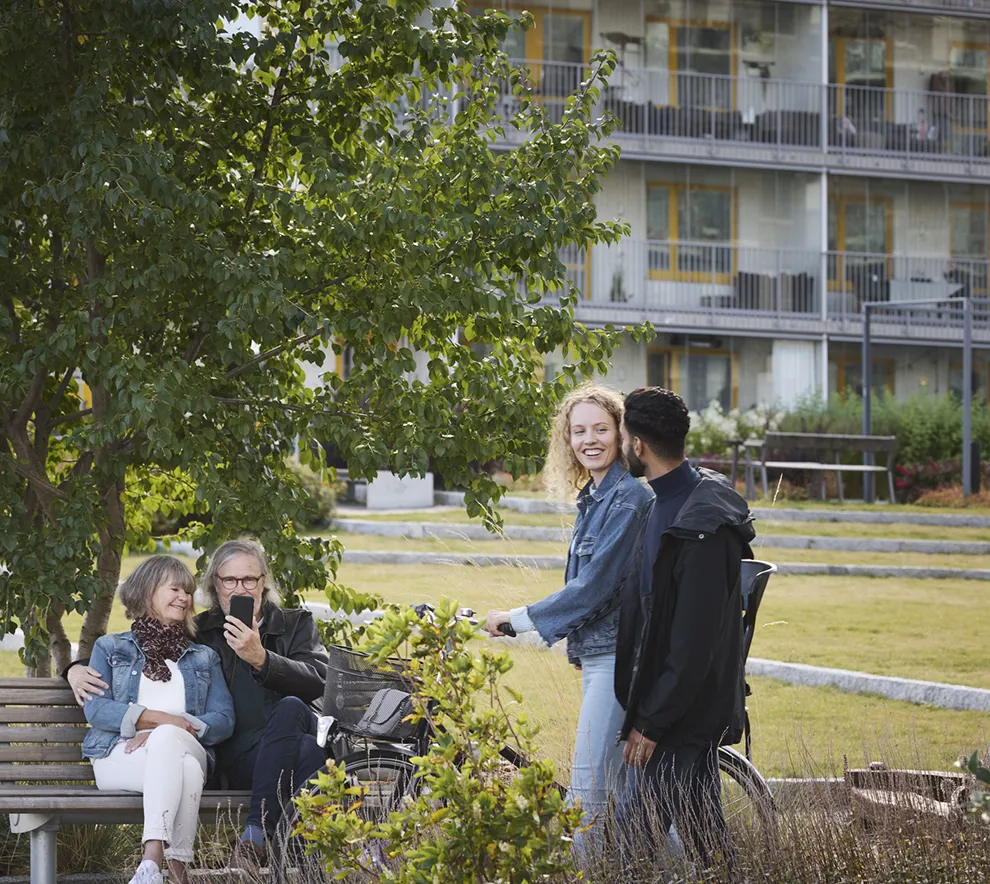 Et eldre par på benk og et ungt par med sykkel i uteområdet til et boligkompleks.