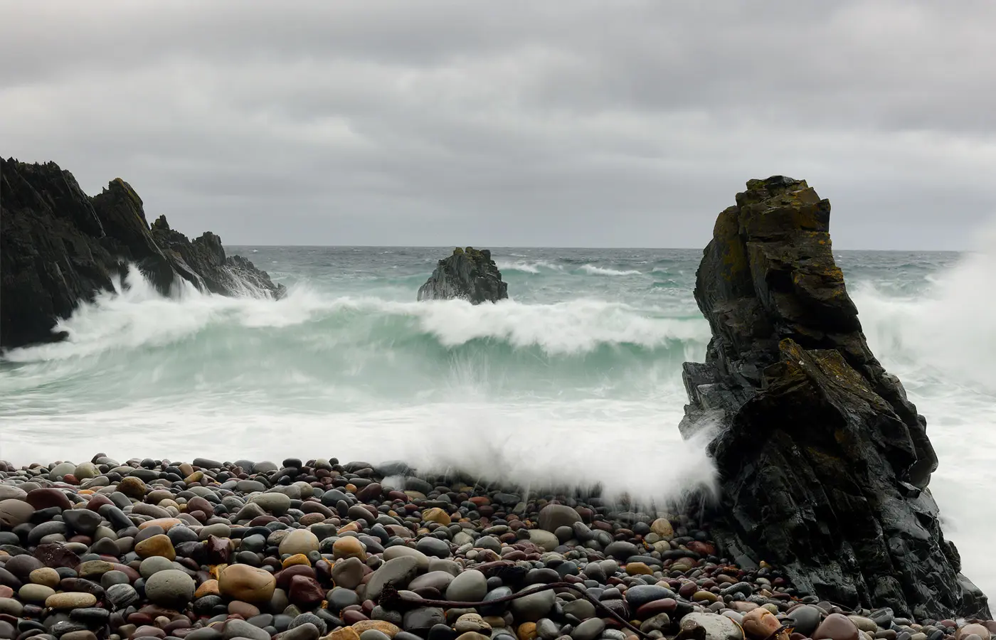 Kraftige bølger slår inn mot mørke klipper på en steinstrand langs en dramatisk kyst under overskyet himmel.