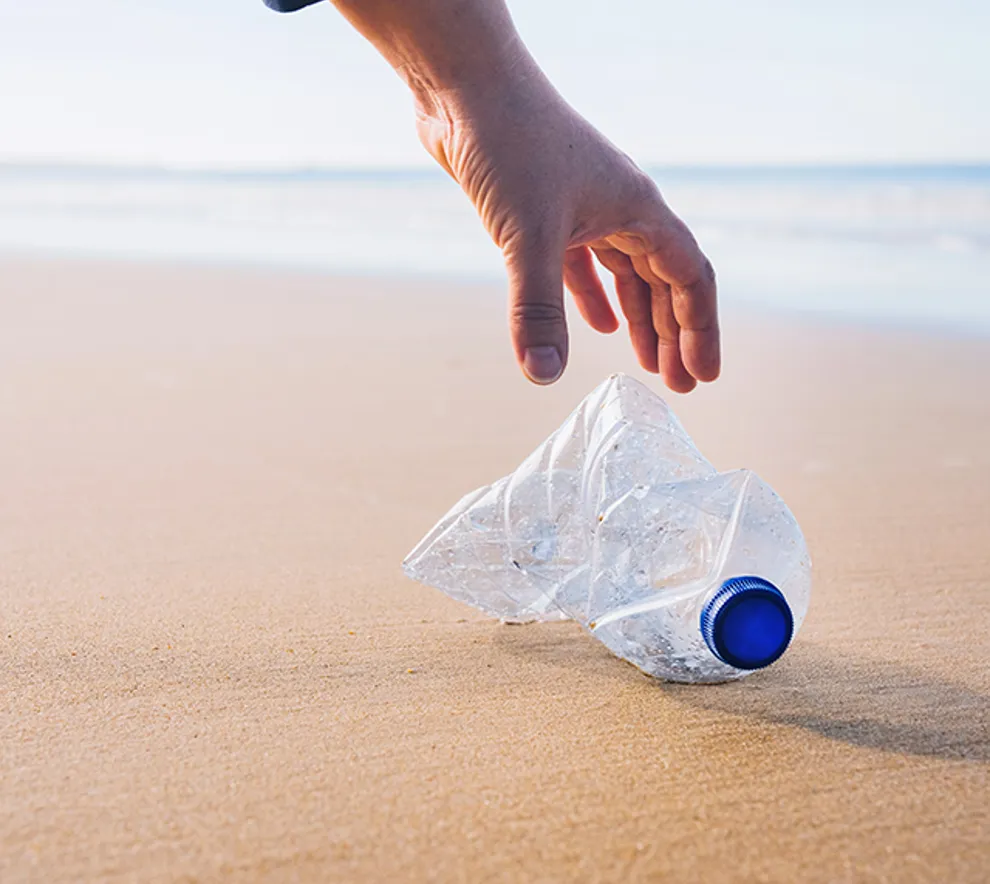 En plastflaske ligger på en fin strand, med havet og en person i bakgrunnen. En hånd rekker ned for å ta opp flasken.