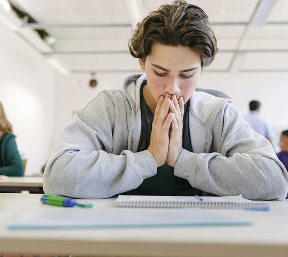 En ung gutt sitter ved pulten sin og virker fortvilt over en prøve eller et valg som må tas.