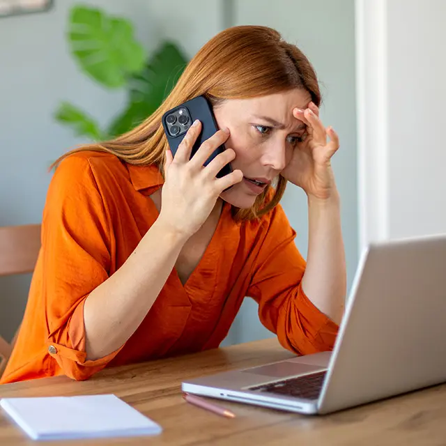 Kvinne sitter ved et bord med pc foran seg, snakker i telefonen