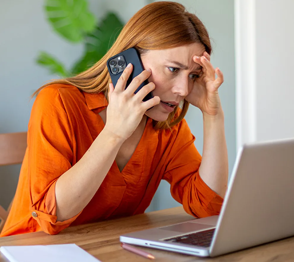 Kvinne sitter ved et bord med pc foran seg, snakker i telefonen