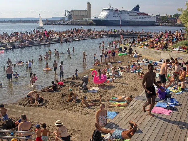 Folksom sommerdag på Sørenga i Oslo – mennesker bader, soler seg og koser seg ved sjøen med cruisebåt i bakgrunnen.