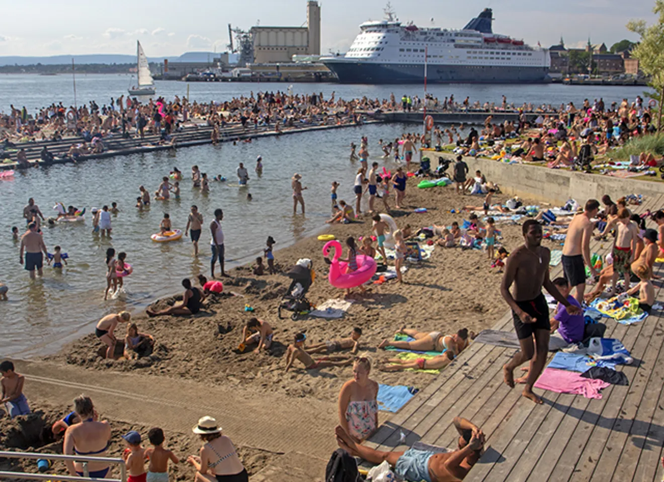 Folksom sommerdag på Sørenga i Oslo – mennesker bader, soler seg og koser seg ved sjøen med cruisebåt i bakgrunnen.