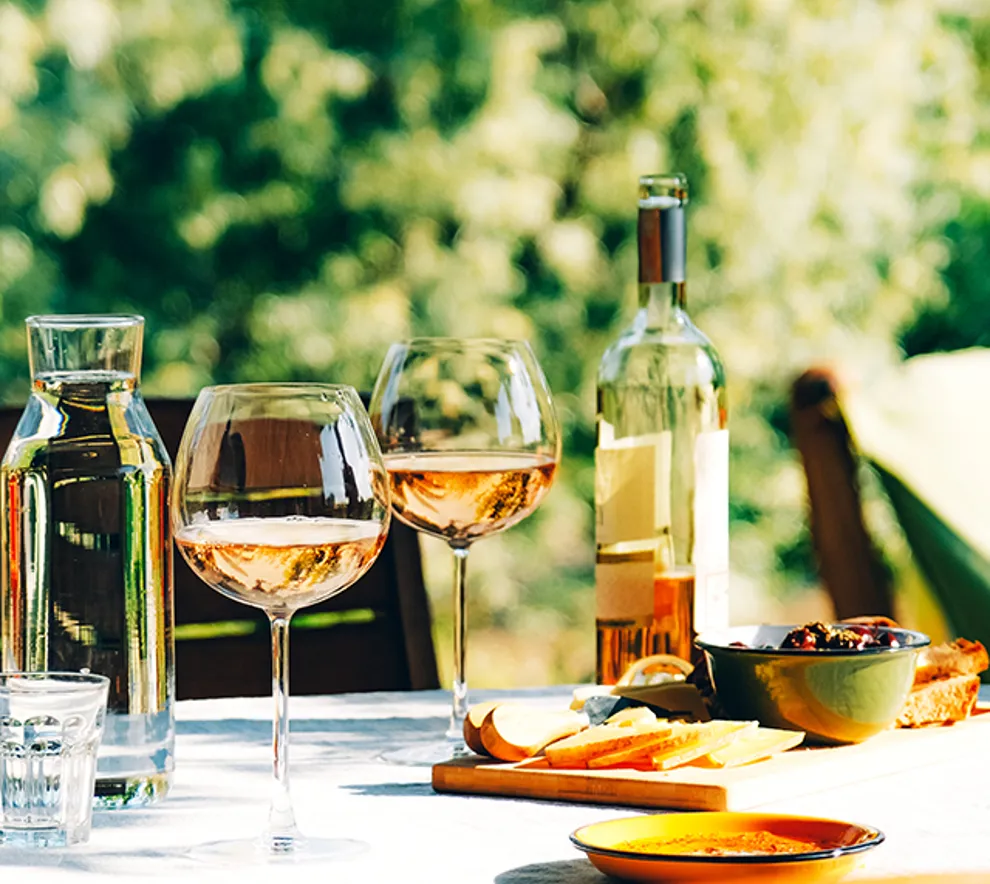To vinglass med rosé på et bord med sommerlige frukter servert rundt. Solen skinner på bordet.