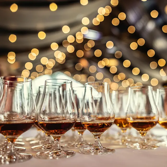 Whiskyglass med gyllen whisky på rekke under en smaking eller festival, med varme lyseffekter i bakgrunnen.