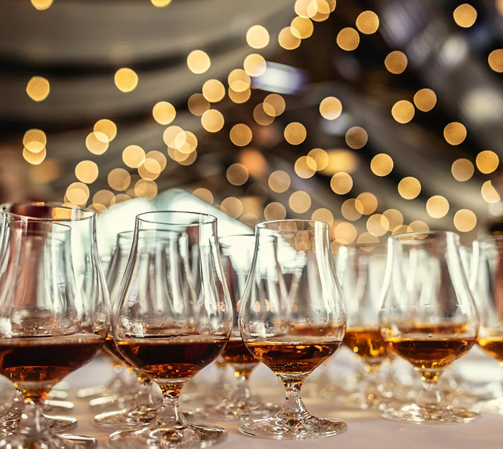 Whiskyglass med gyllen whisky på rekke under en smaking eller festival, med varme lyseffekter i bakgrunnen.