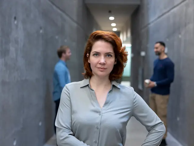 Illustrasjonsbilde av en ung kvinne stående i en gang foran to menn og smiler til kamera