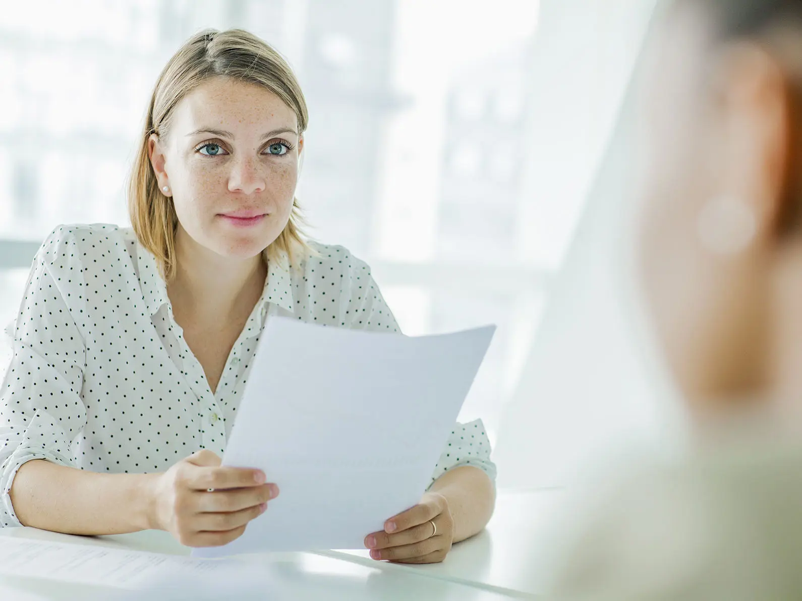En ung kvinne sitter ved et bord og holder flere ark med papir mens hun ser på en annen person utenfor bildet