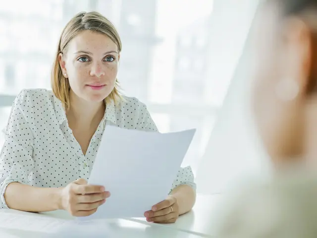 En ung kvinne sitter ved et bord og holder flere ark med papir mens hun ser på en annen person utenfor bildet