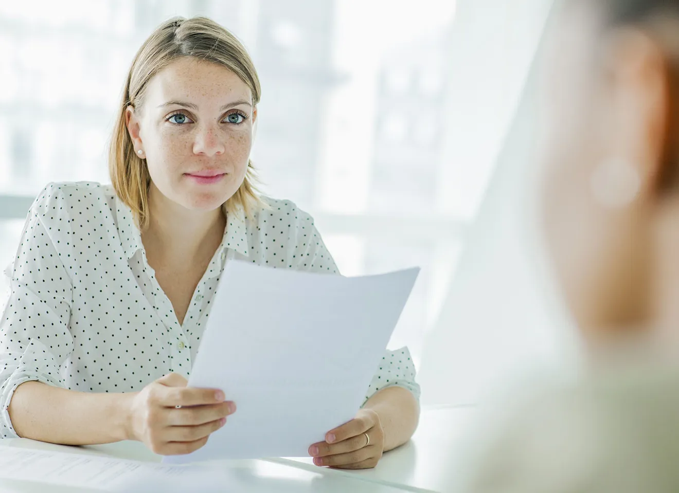 En ung kvinne sitter ved et bord og holder flere ark med papir mens hun ser på en annen person utenfor bildet