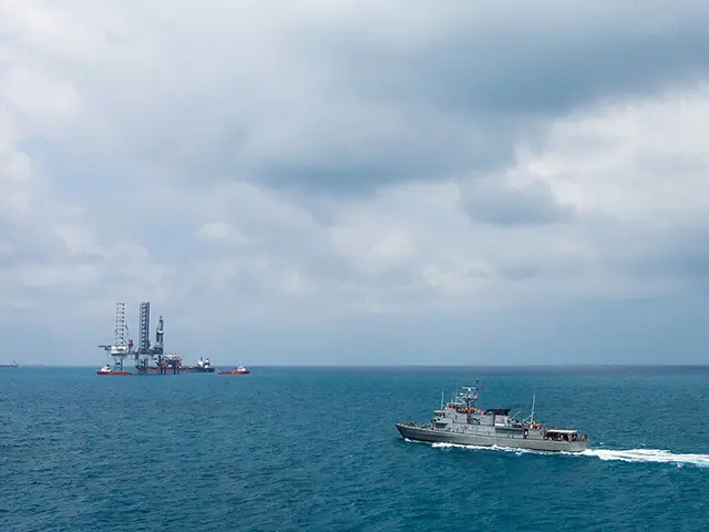 Et krigsskip i forgrunnen patruljerer rundt en oljerigg som skimtes i bakgrunnen.