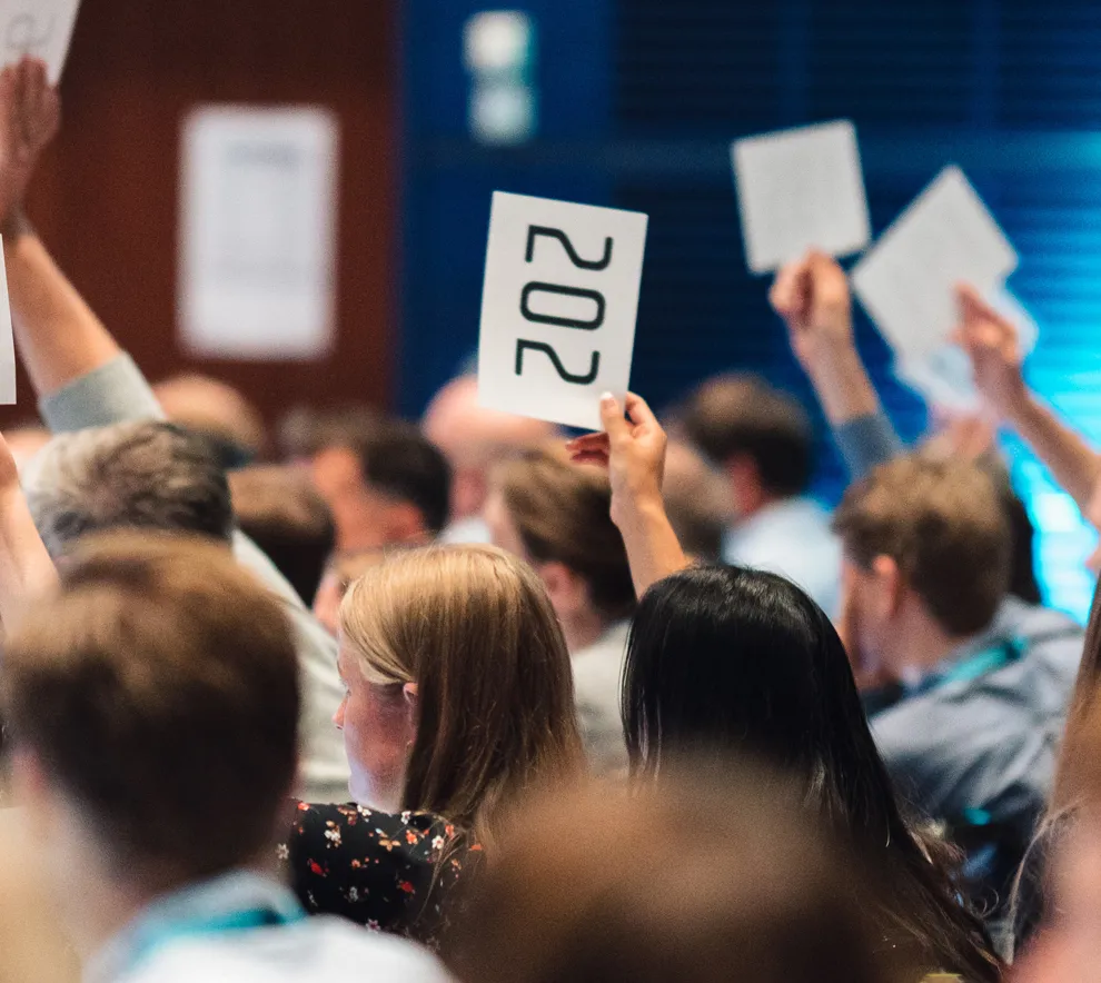 En stor gruppe mennesker sitter i en stor sal og mange rekker opp hender for å stemme for noe