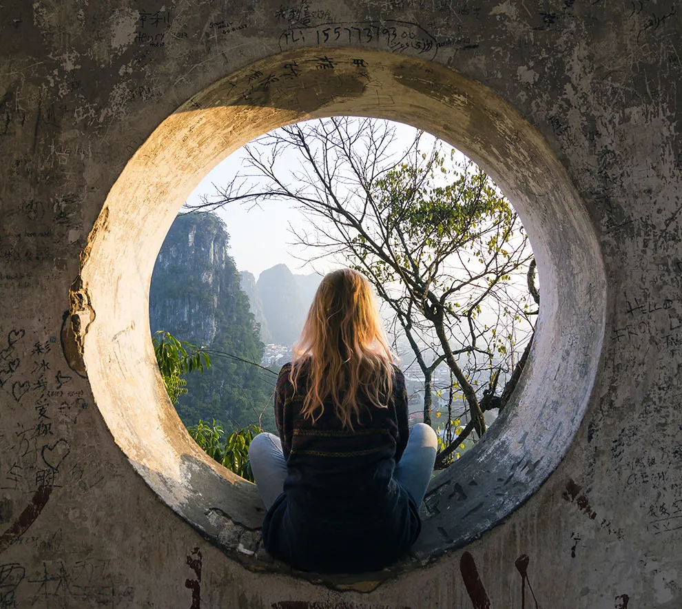 Kvinne sitter i en sirkelformet steinstatue og stirrer ut over et landskap med ryggen til