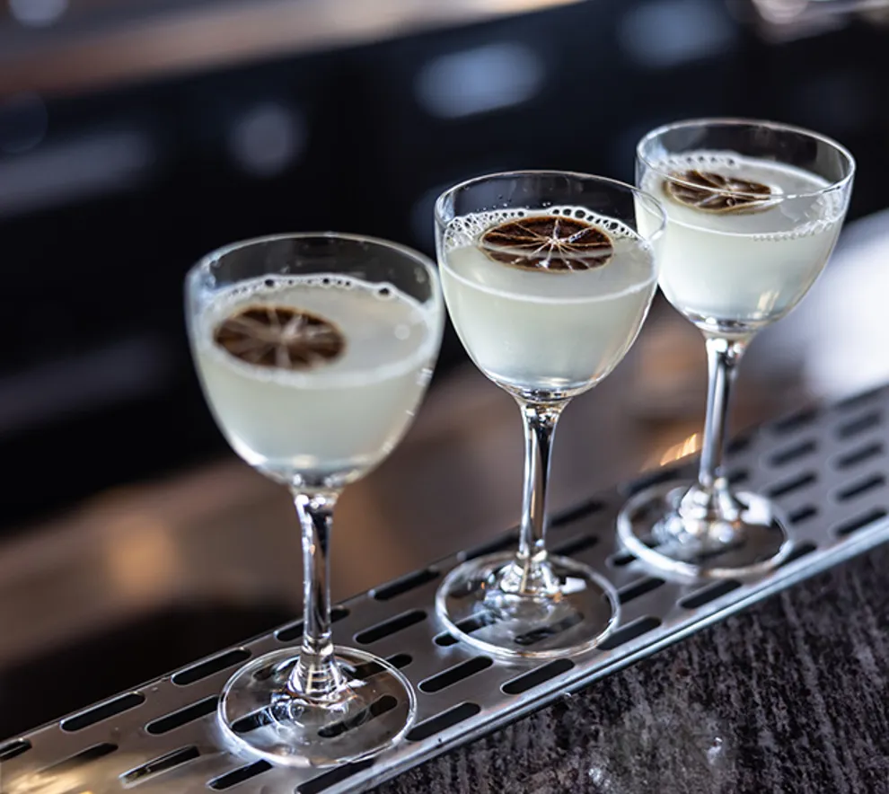Tre elegante cocktails med skum på toppen og tørket sitrusgarnityr, servert i coupeglass på en stilig bardisk.