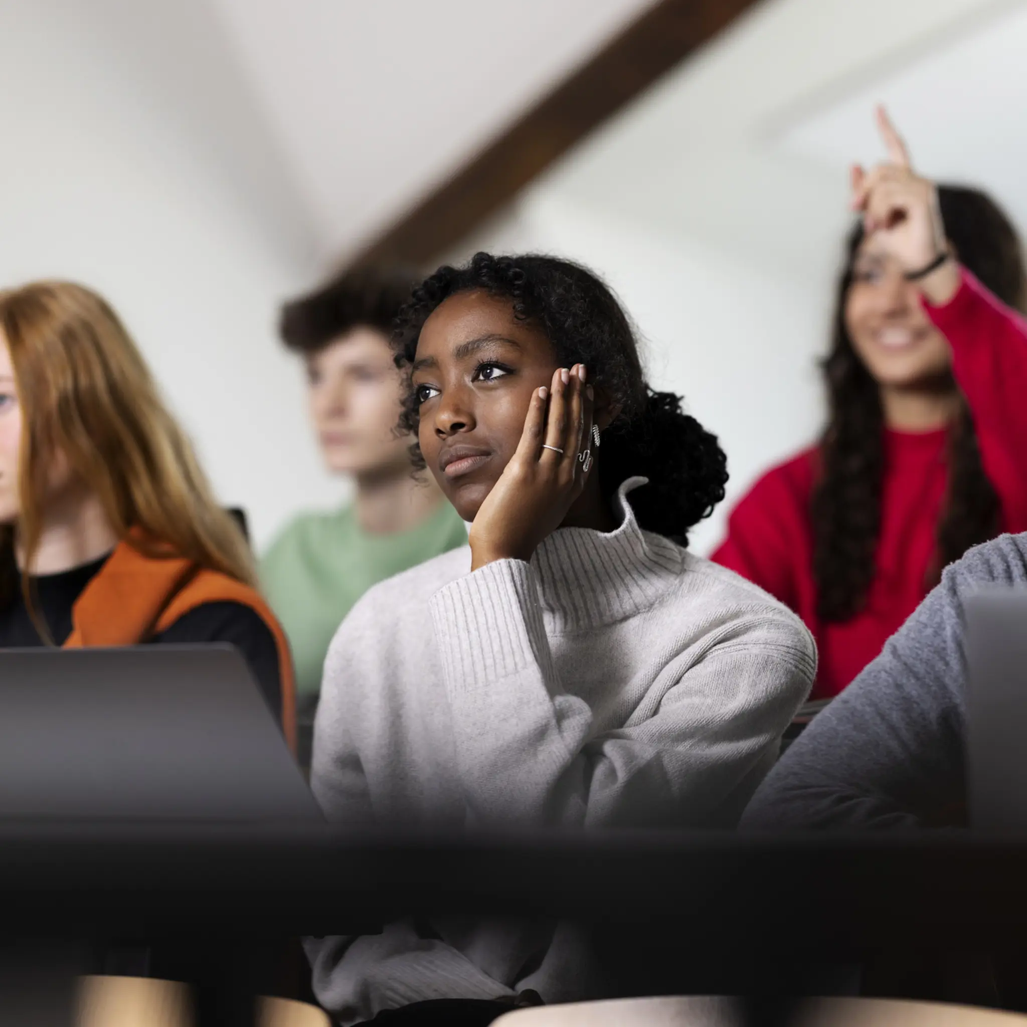 Nærbilde av noen studenter med forskjellige hudfarger, en som sitte og lytter og en som rekker opp en hånd
