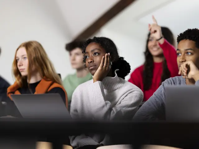 Nærbilde av noen studenter med forskjellige hudfarger, en som sitte og lytter og en som rekker opp en hånd