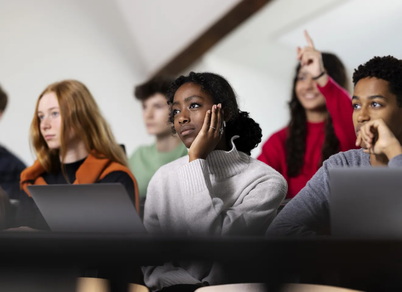 Nærbilde av noen studenter med forskjellige hudfarger, en som sitte og lytter og en som rekker opp en hånd
